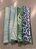 Three folded outdoor rugs showing different color and pattern designs.
