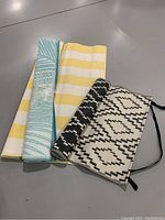 Three rolled outdoor rugs on a flat surface, showing colors and patterns: yellow and white stripes, turquoise geometric, black and white geometric with ties.