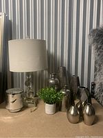 Full shot showing the table lamp, green faux plant, and multiple metallic decorative vases in varying shapes and finishes on a brown surface with corrugated metal background.