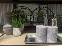Artificial green plants in various ceramic pots along with two gray candles on a metal tray and two silver soap dispensers on a shelf