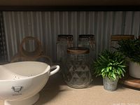 Photo showing kitchen containers including a large white colander, glass jars with wooden lids, small turntable, and faux greenery.