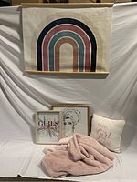 Photo showing fabric wall hanging with rainbow design, pink plush blanket, two framed artworks, and decorative cushion