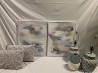 Two abstract wall art pieces positioned side-by-side with two gray textured decorative cushions on left and two porcelain lamps (no shades) on right with a small decorative porcelain piece in front