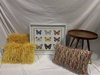 Full decor set showing framed butterfly print art, yellow fluffy cushions, multicolor patterned cushion, and round wooden table on white backdrop.
