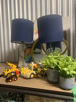 Two blue fabric shade table lamps, round mirror with rope detailing, colorful framed art, faux greenery, and several children's toys on wood surface.