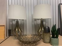Two brass-tone metal table lamps with white fabric shades with a decorative bowl, vase, and potted plant on the table.