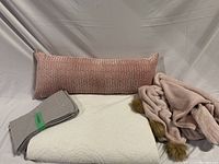Photo showing pink lombar cushion, folded grey pillow case set, white quilt, and pink plush blanket with brown pom-poms.