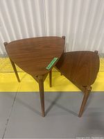 Photo showing two wooden nesting tables stacked, visible triangular tops with wood grain and rounded legs, yellow and gray floor background.