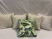 Seven outdoor cushions arranged in two rows showing geometric beige patterned cushions and one tropical leaf cushion.