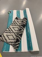 Two rolled outdoor rugs side by side on floor; larger turquoise and white striped rug with tag, smaller with black and white geometric pattern.