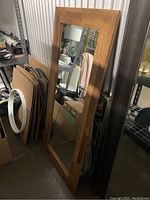Full view of the tall rectangular mirror with a wood frame leaning against a wall in a storage setting.
