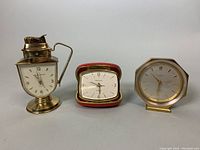 Three vintage traveller clocks including a brass teapot lighter clock, a red leather folding clock, and a hexagonal brass alarm clock.