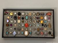 Tray of over 60 assorted costume rings laid out on padded display surface. Varied metal base colors primarily silver and gold-tone with multiple faux stones in different shapes and colors. Some rings have missing stones.