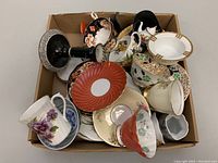 Box full of assorted vintage teacups, saucers, miniatures, plates in various colors and designs, some with visible wear and damage