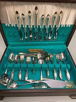 Top view of the flatware set inside the open wooden chest, showing various forks, spoons, and serving utensils neatly arranged in turquoise felt slots.