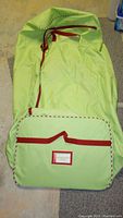 Light green fabric Christmas tree storage box on floor, showing overall shape with red straps and attached front pocket with label holder.