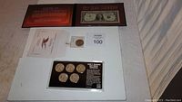 Lot arranged on white surface showing coin collection folder, five Kennedy half dollar coins in black holder, 1957 Silver Certificate bill in folder, single coin in plastic holder and related documents.