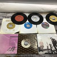 Overview of 7 45 RPM vinyl records laid out with some paper sleeves, showing various record labels and colors.