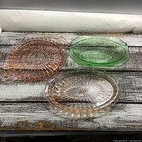 Three carnival glass pieces on a rustic wooden surface including a round pink plate, an oval pink plate, and a green oval dish.