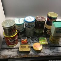 Photo showing multiple tobacco tins arranged in rows on wooden surface, with various brands and sizes visible.