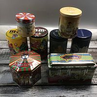 Nine assorted M&M's and Hershey collector tins grouped on a rustic wood surface, various shapes including round, octagonal, and rectangular, colorful and detailed designs