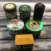 Six assorted collectible tins arranged on a wooden surface, showcasing varied designs including nature scenes, vintage portraits, and brand labels