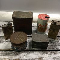 Six vintage tins positioned on a wooden surface including tea, coffee, oil, fly fluid, and kerol tins.