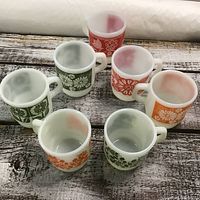 Seven Fire King coffee mugs with flower motifs in green, red, and orange on a wooden surface.