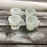 Four white Fire King mugs on wood plank surface. One mug has a flower design on one side.