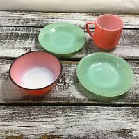 Set of 2 green jadeite glass saucers, 1 small pinkish bowl, and 1 pink mug arranged on rustic wood surface.