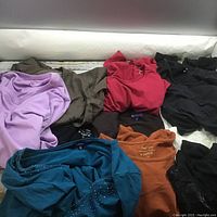 Assorted ladies sweaters and tops in various colors including lavender, brown, red, black, teal with embellishments, and orange, laid out showing fabric and texture.