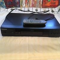 Front view of Panasonic black DVD player with remote on top, showing branding and disc tray.