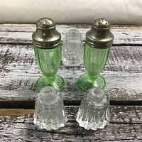Front view of two green depression glass salt and pepper shakers with metal tops, and two clear cut glass shaker bases with no tops shown on rustic wooden surface.