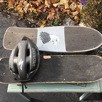 Two skateboards stacked with black Specialized helmet in front, placed on a small table outside on an asphalt surface with fallen leaves nearby.
