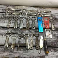 Full view of all 23 bottle openers spread on wooden surface showing various metal and plastic designs and shapes