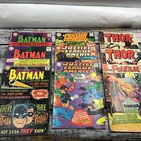 Photo showing full lot of 9 comic books including Batman, Justice League of America, Thor, and The Phantom comics arranged on white surface with wood pattern background.