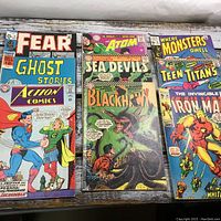 Collection of vintage comics grouped together on a wooden surface showing titles Fear, Ghost Stories, Action Comics, The Atom, Sea Devils, Blackhawk, Where Monsters Dwell, Teen Titans, and Iron Man.