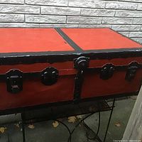 Vintage trunk with red panels and black metal hardware set against a light brick wall background, shown from slightly different angles