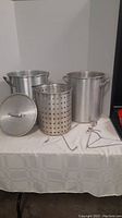 Two large aluminum deep fryer pots with handles, a lid, perforated strainer basket, and metal accessory hooks on a table.