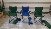 Three camping chairs arranged side by side, two green and one blue, with matching carry bags placed in front of each chair. Image shows overall condition including visible minor damages.