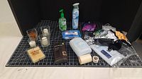 Photo showing various bathroom miscellaneous items arranged on black and white grid surface, including candles, soap, hand sanitizer, air freshener, wipes, container, toiletry bag, toilet paper, tape, and small accessory bags.