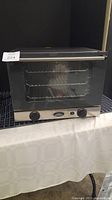 Full view of Cadco Unox OV-250 convection oven showing front glass door, three internal shelves, temperature knobs, and stainless steel body on a black grid table with a white cloth beneath.