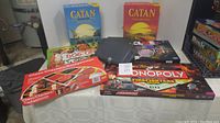 Seven assorted board games displayed on table, including multiple Catan editions, Monopoly Firefighters edition, Clue, Dog-Opoly, Chess/Checkers/Backgammon set and one black box game.