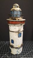 Front view of handmade metal lighthouse showing distressed white paint with blue windows and rust-colored band around the top.