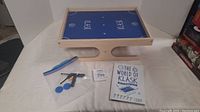 Wooden Klask game board with accessories including magnetic strikers, blue pieces and instruction manual on white background.