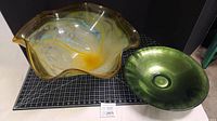 Two large display bowls on black grid surface with measurement scale visible. The colorful scalloped edge bowl is larger, about 21 inches wide, with multi-color swirled design. The green bowl is round, about 16 inches diameter, thick glass with a smooth green finish.