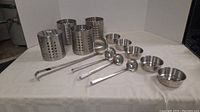 Photo showing 4 cylindrical utensil holders, 5 small metal bowls and 4 metal ladles arranged on a table.