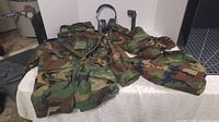 Camouflage jacket, pants, headphones, flashlight, and gear displayed on table