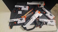 Front angled view of six Nerf Rival blasters arranged on a black grid surface, showing white and black bodies with orange triggers and accents, along with two transparent speed loaders filled with orange foam balls and loose balls scattered.