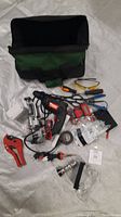 Tool bag opened with tools spread out including Drillmaster drill, soldering iron, pipe cutter, safety glasses, pliers, wire cutters, electrical tape and other items.